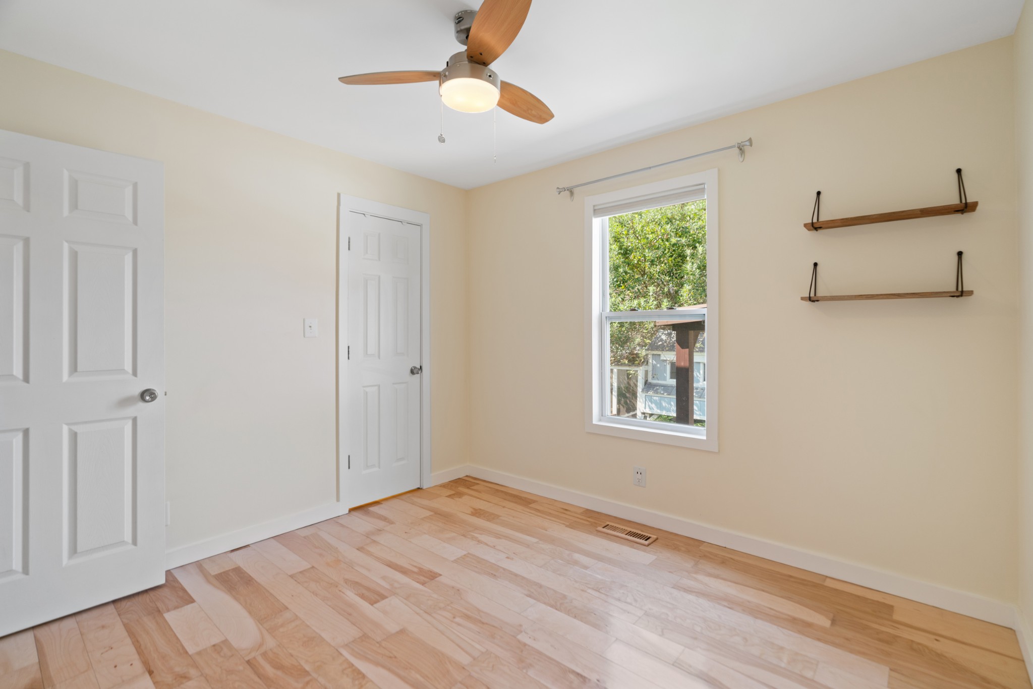 Empty bedroom with pale wood flooring, a ceiling fan, window, and wall shelves.