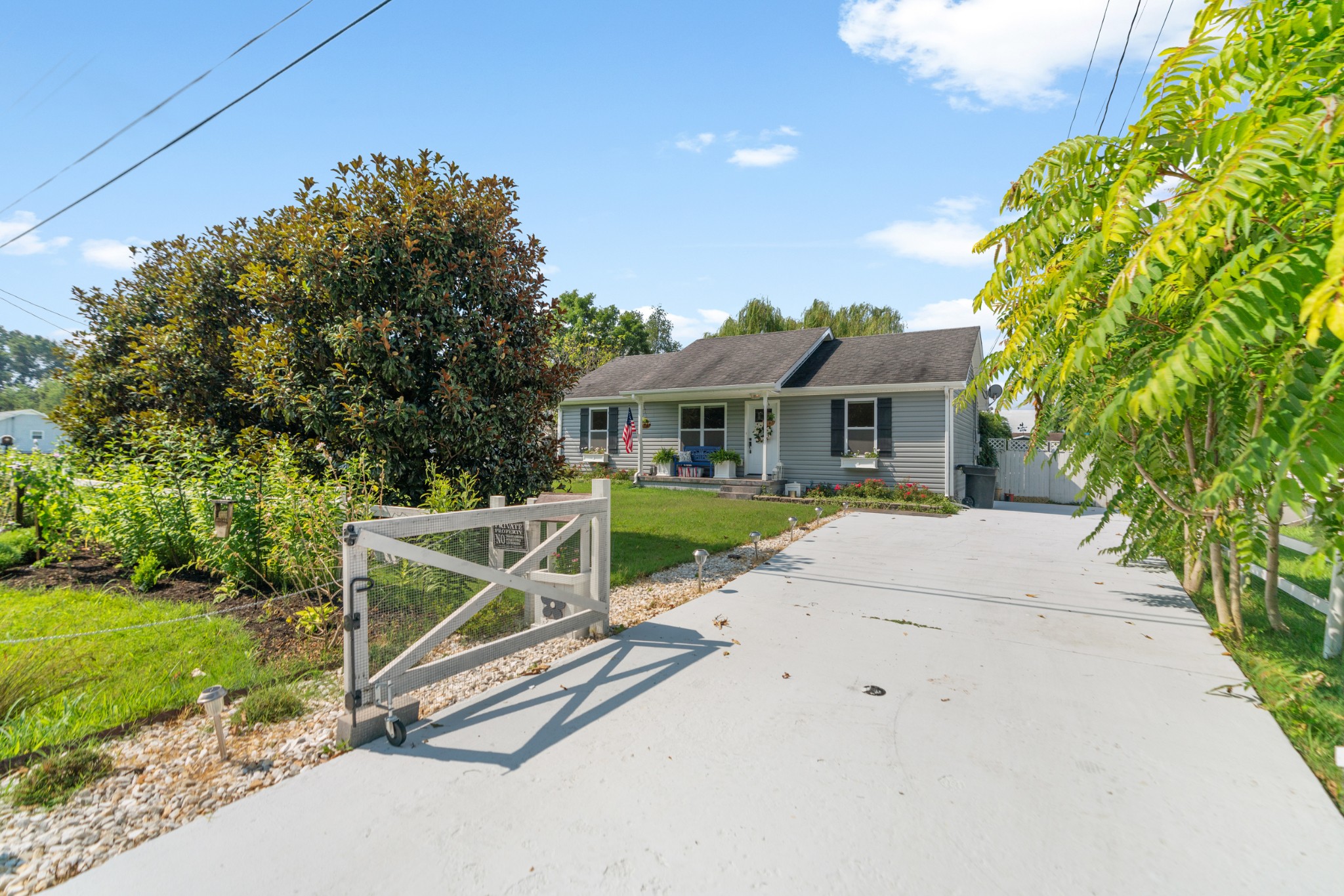 Gray single-story home with a covered porch, broad driveway, open gate, and landscaped garden.