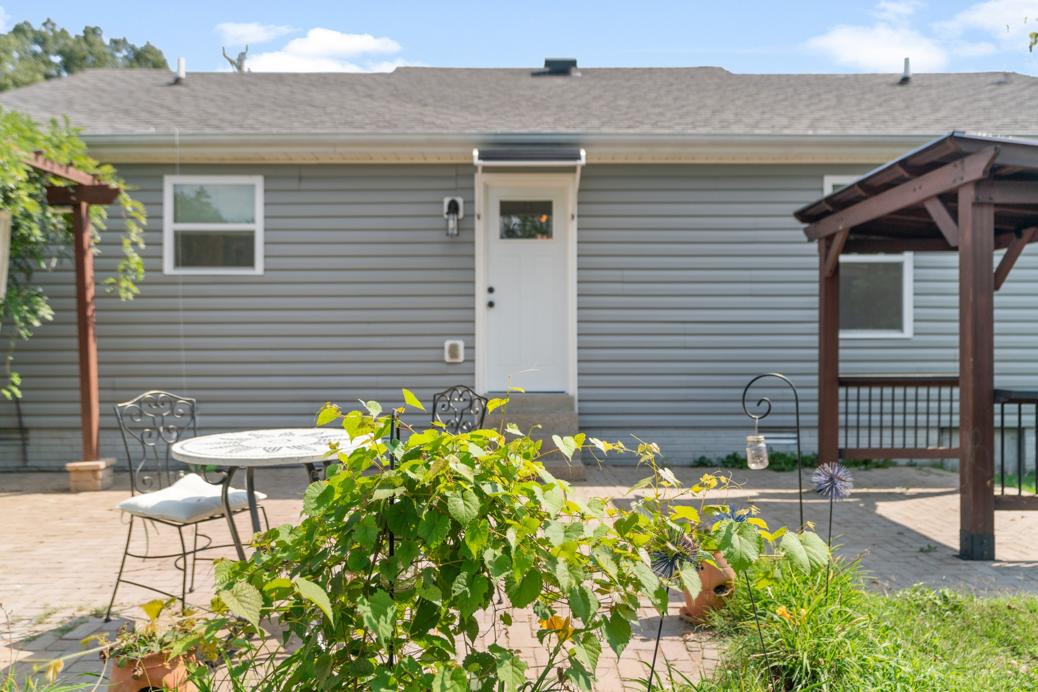 Home’s rear patio with a wooden pergola, metal table and chairs, and leafy garden plants.