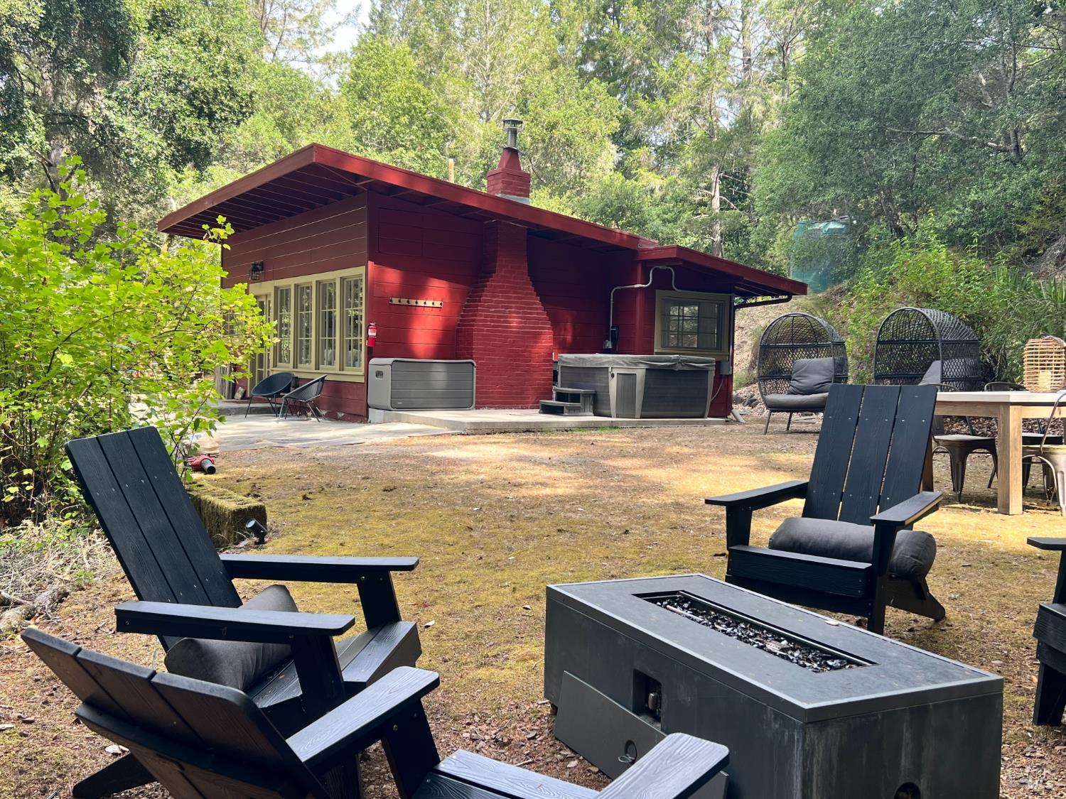 Red modern home in the woods with a furnished patio, hot tubs, and a rectangular fire pit.