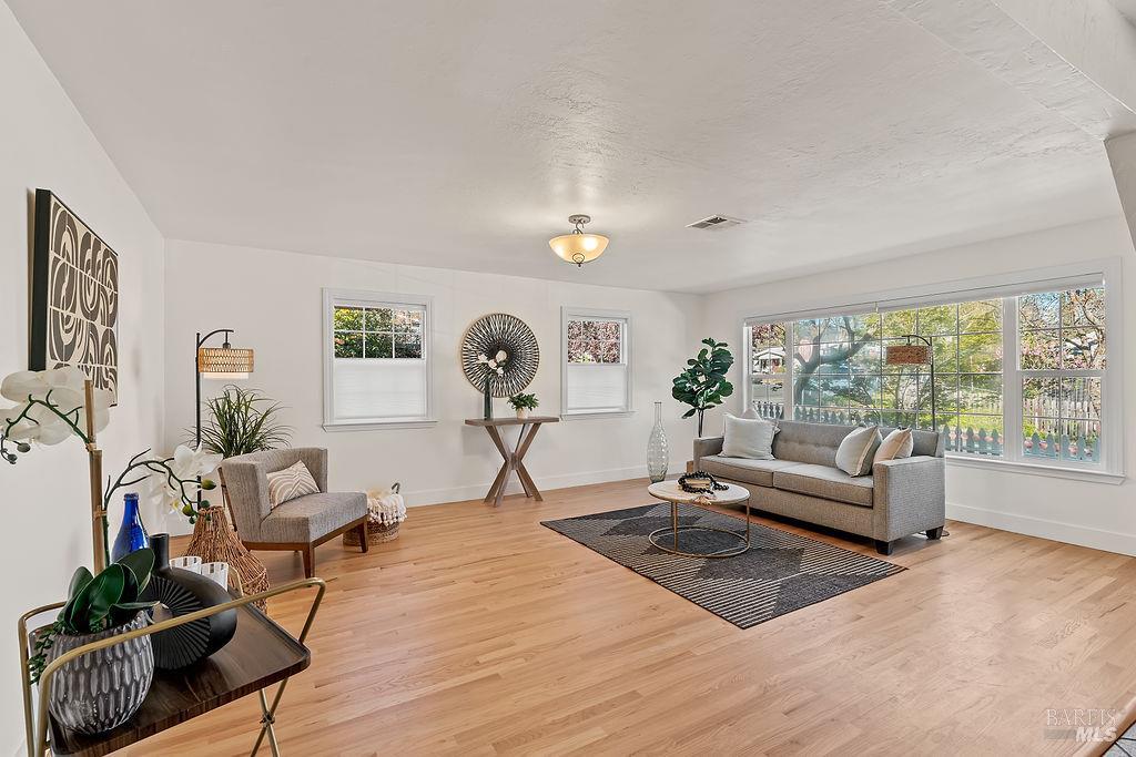 Staged living room with hardwood floors, gray sofa, accent chairs, and large windows overlooking a fenced yard.