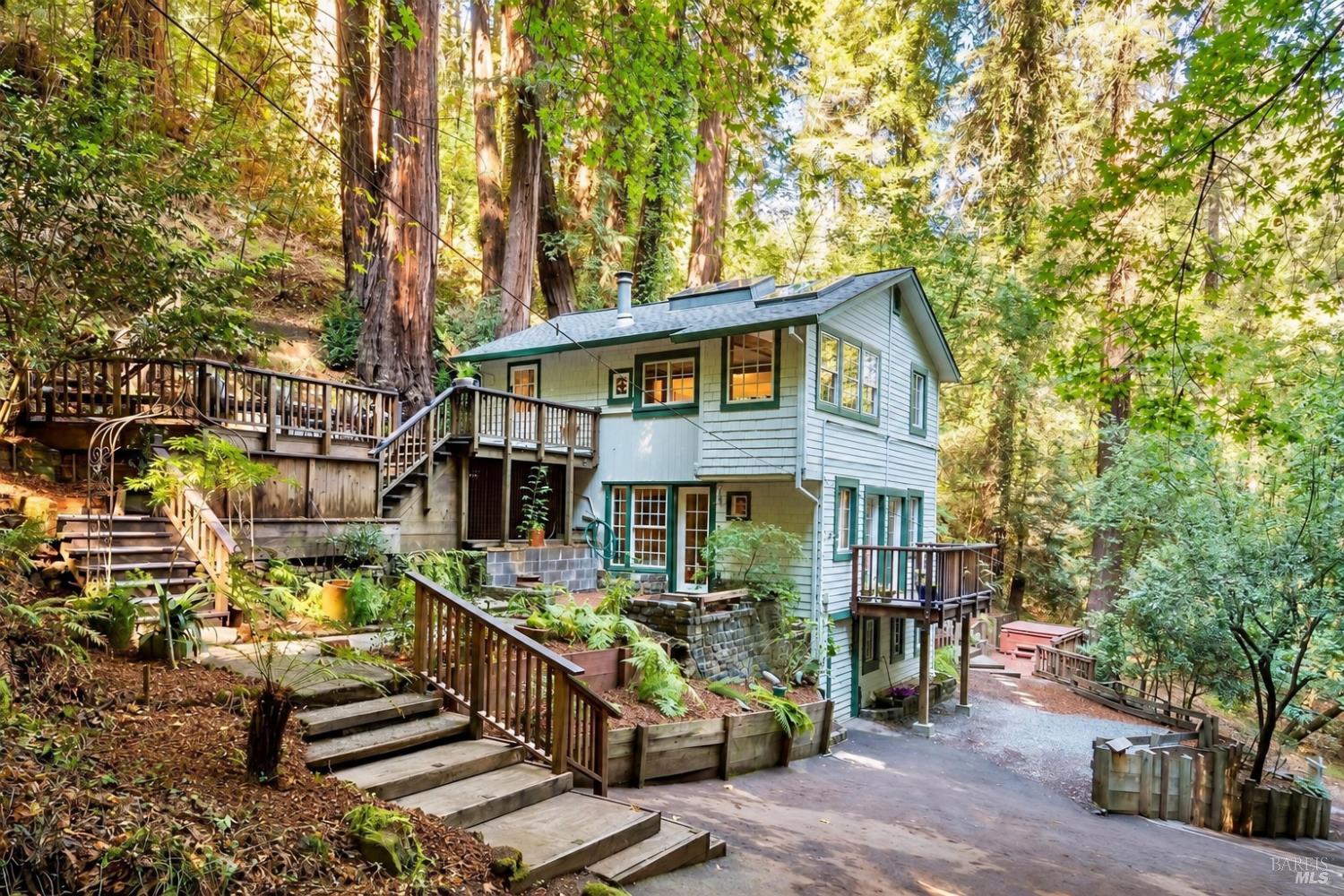 Light-blue two-story home nestled among redwoods, with expansive wooden decks and hillside stairs.