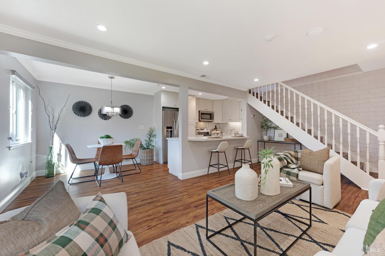 Open-plan living room with hardwood floors, neutral furnishings, and a white staircase beside the kitchen.