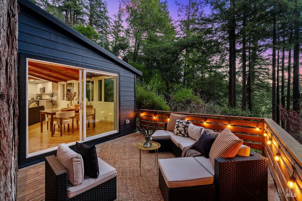 Furnished deck with outdoor seating beside a modern home, surrounded by tall trees at dusk.