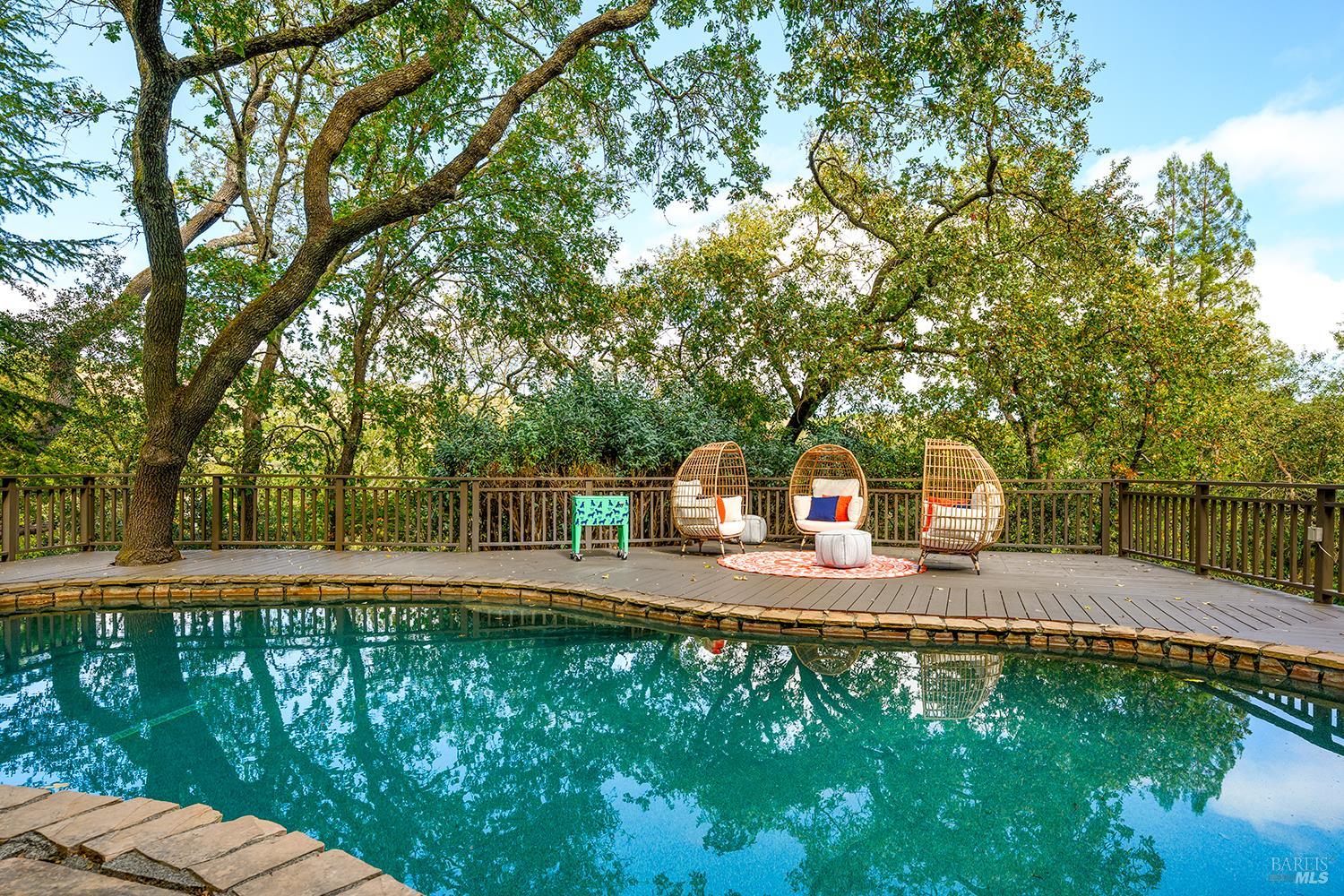 Swimming pool beside a wooden deck with wicker lounge chairs and mature trees.