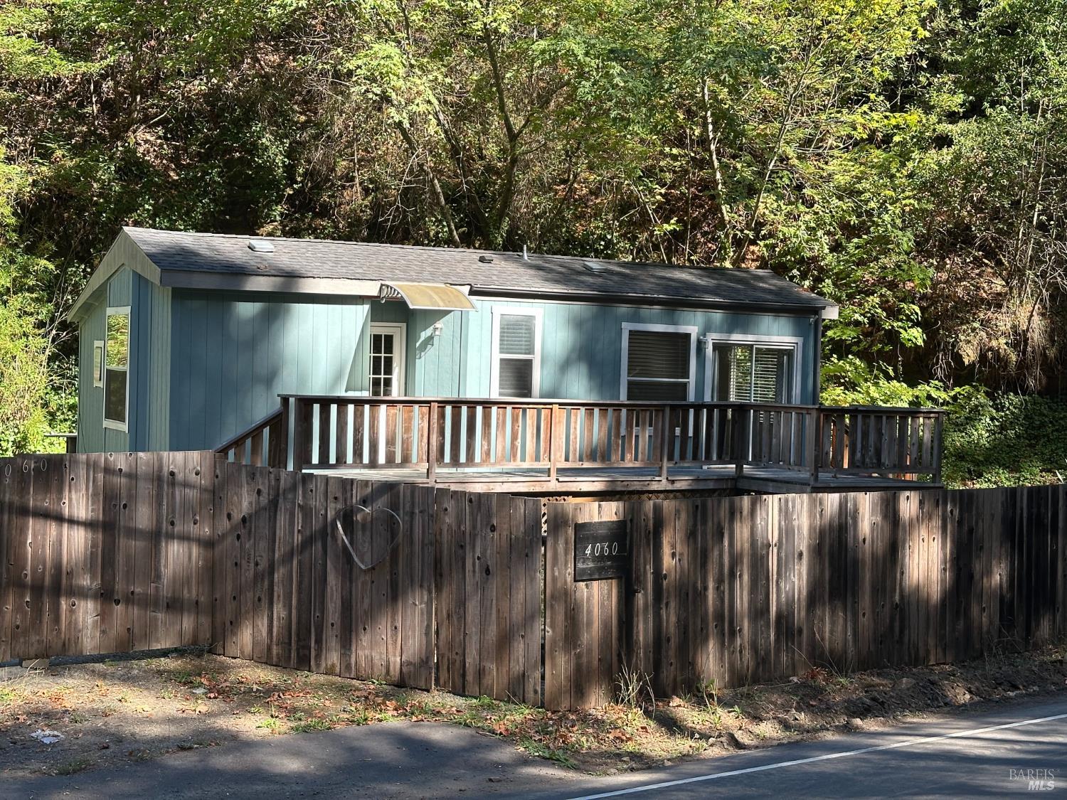 Blue cottage with a long wooden deck behind a tall fence, surrounded by trees.