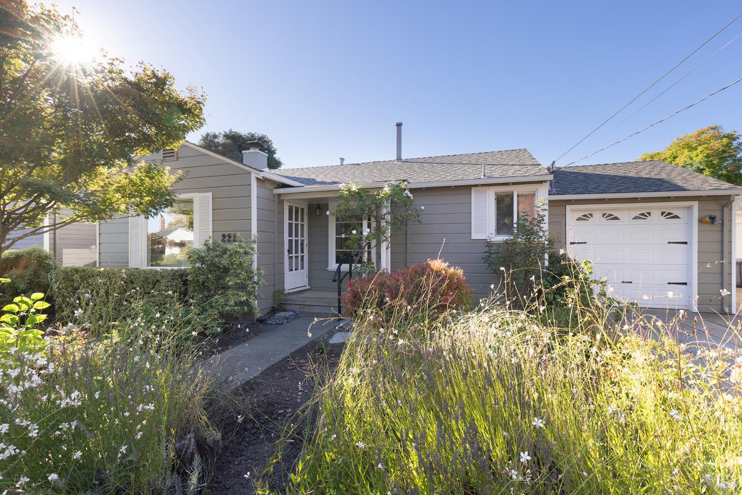 Gray single-story home with white garage door and a sunlit, flower-filled front garden.