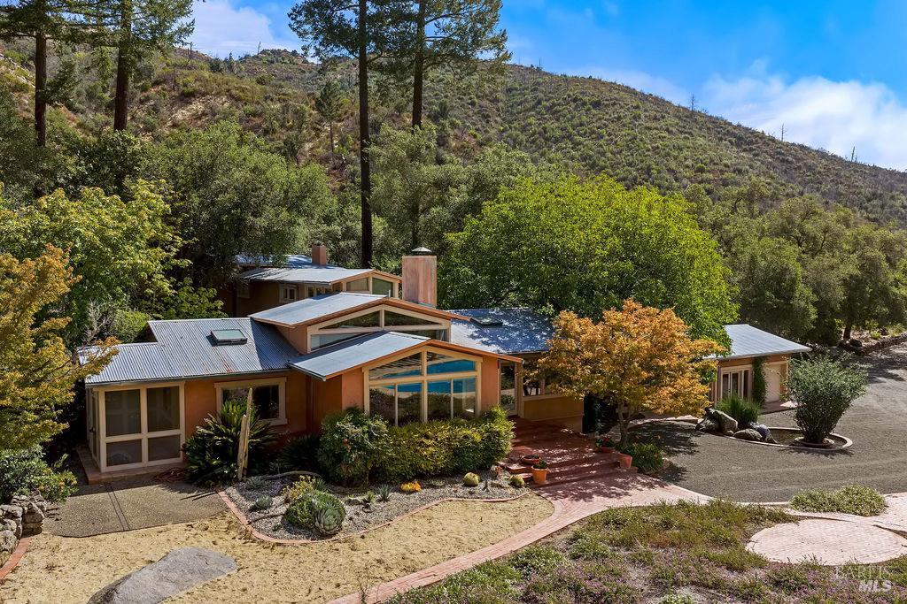 Low modern home with metal roofs, large windows, and landscaped grounds against a wooded hillside.