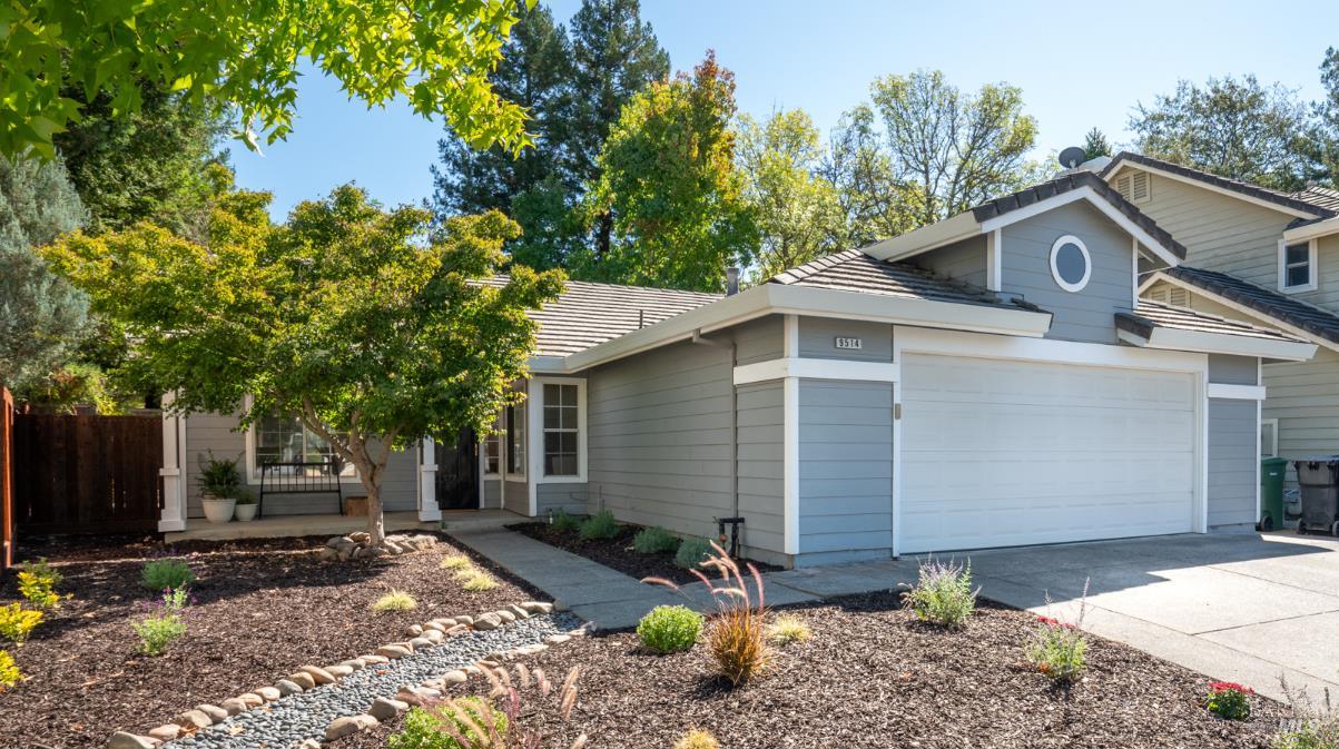 Gray home with white double garage door, landscaped garden beds, and mature trees.
