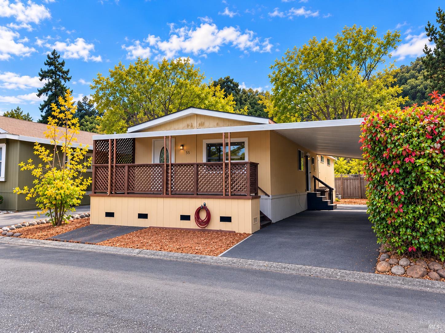Tan manufactured home with a covered lattice porch, attached carport, and landscaped front yard.