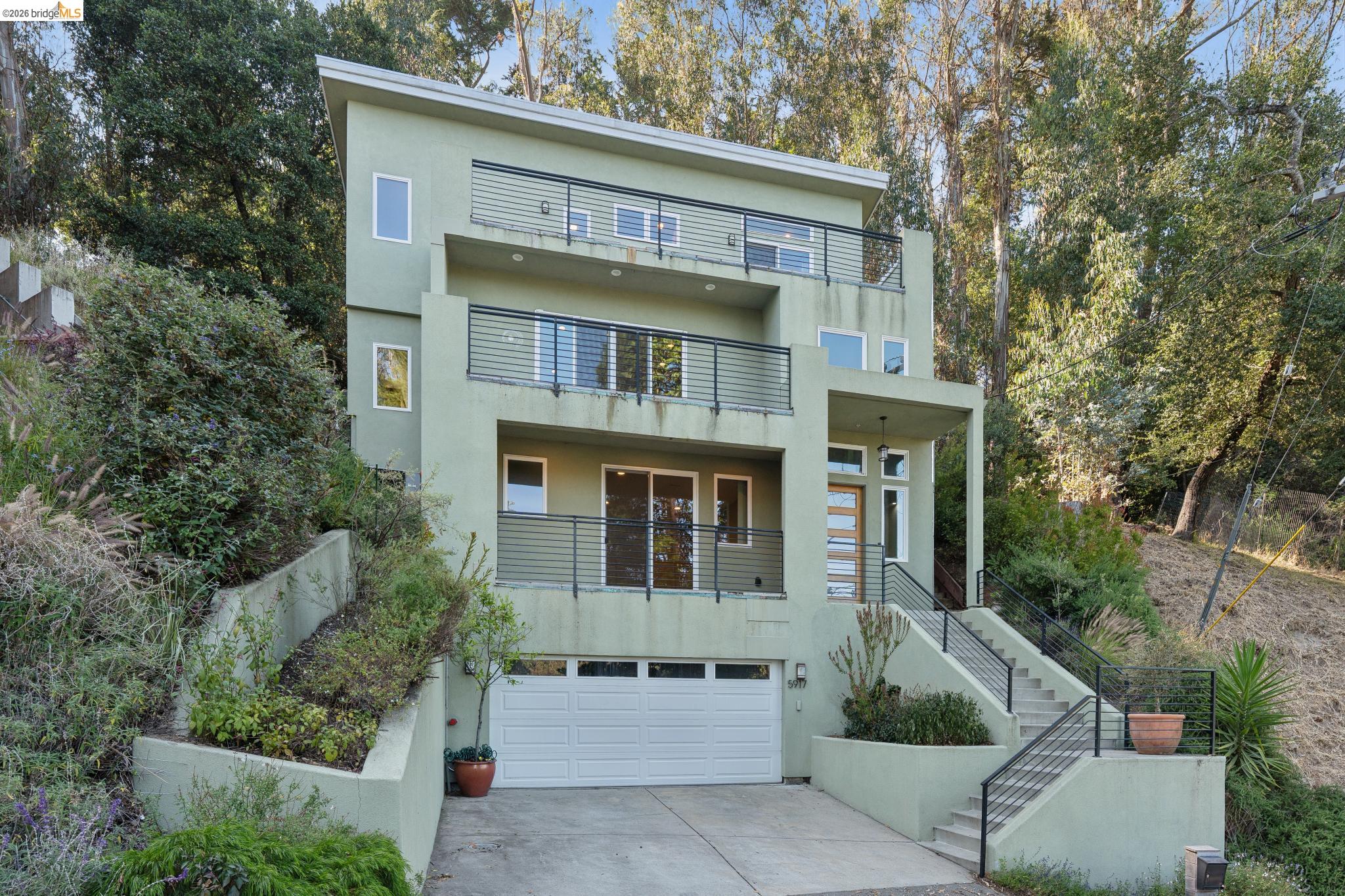 Modern multi-story home with black-railed balconies, garage, and hillside staircase.