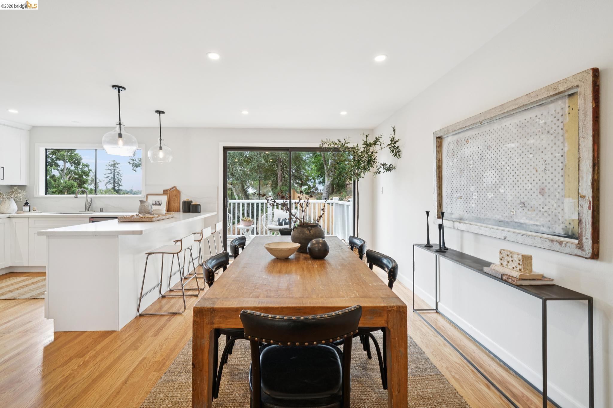 Open-plan dining room with a long wooden table, white kitchen island, and glass doors to a tree-lined balcony.