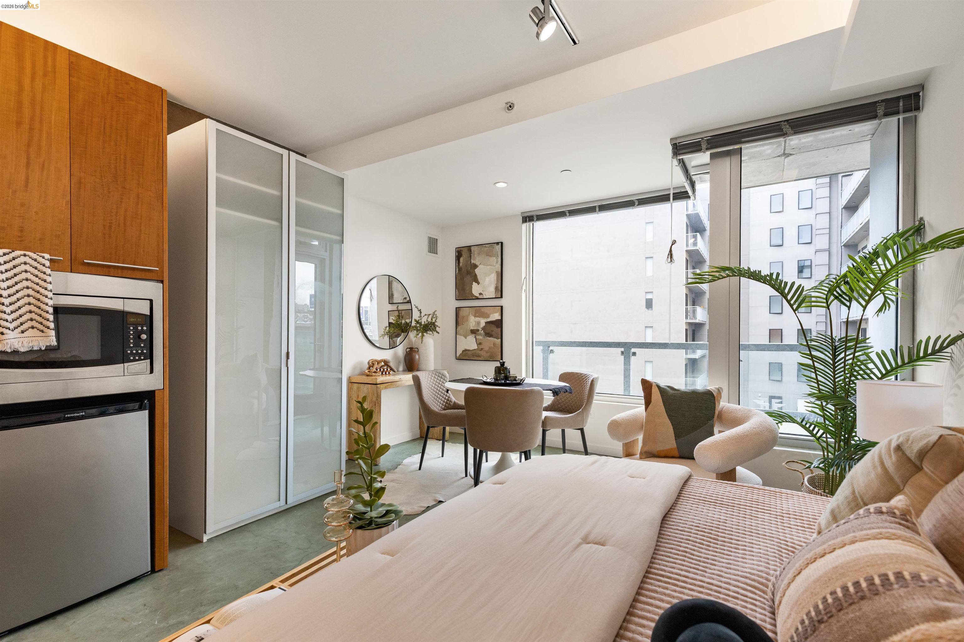 Modern studio living area with wood cabinetry, dining table, and large windows facing nearby buildings.