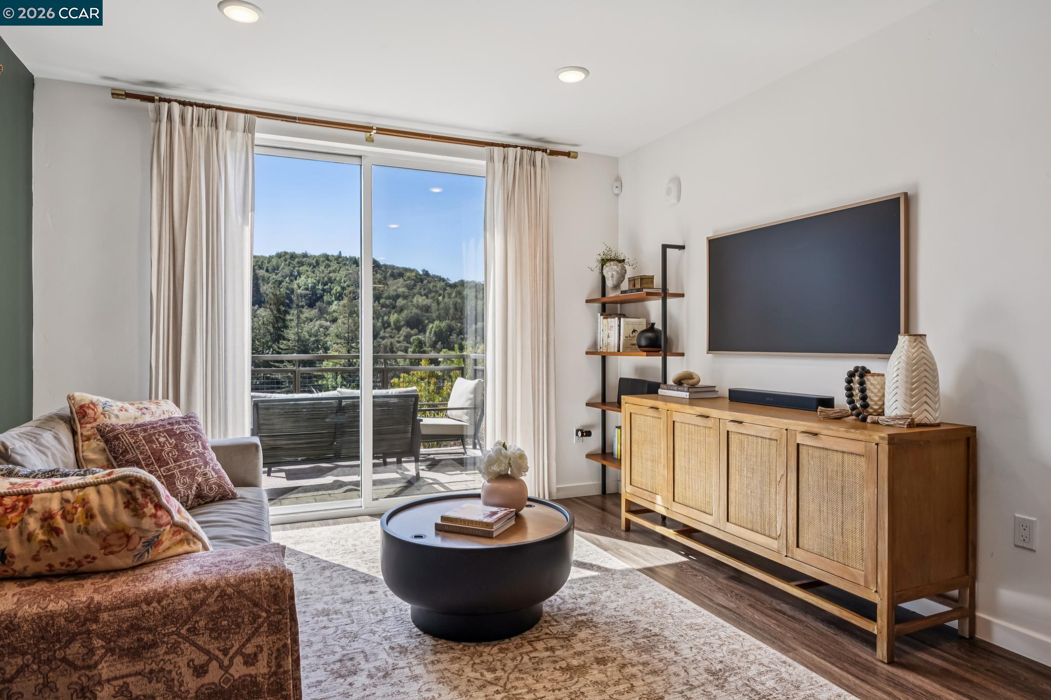 Living room with a balcony overlooking wooded hills, furnished with a sofa and wooden media console.
