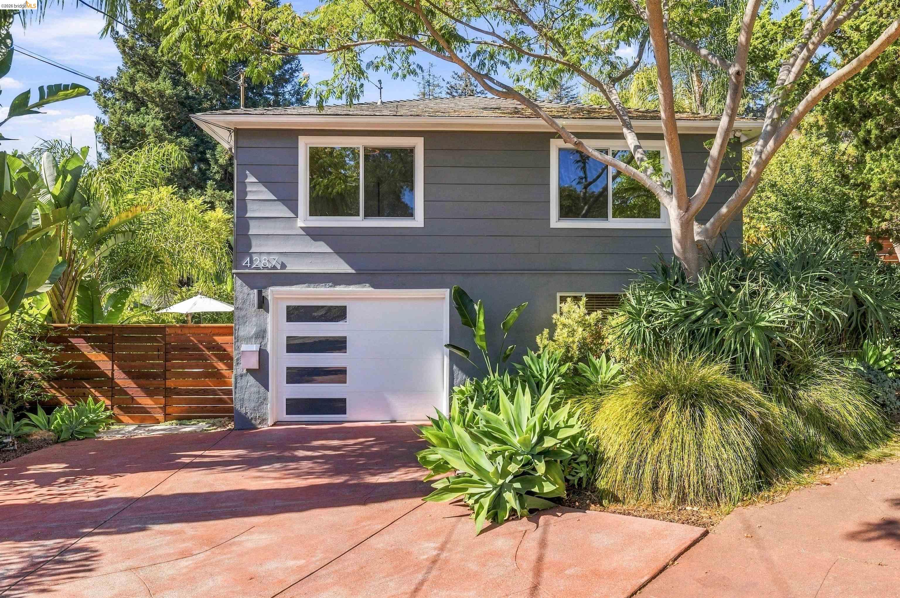 Gray two-story home with a white garage, reddish driveway, and lush tropical landscaping.