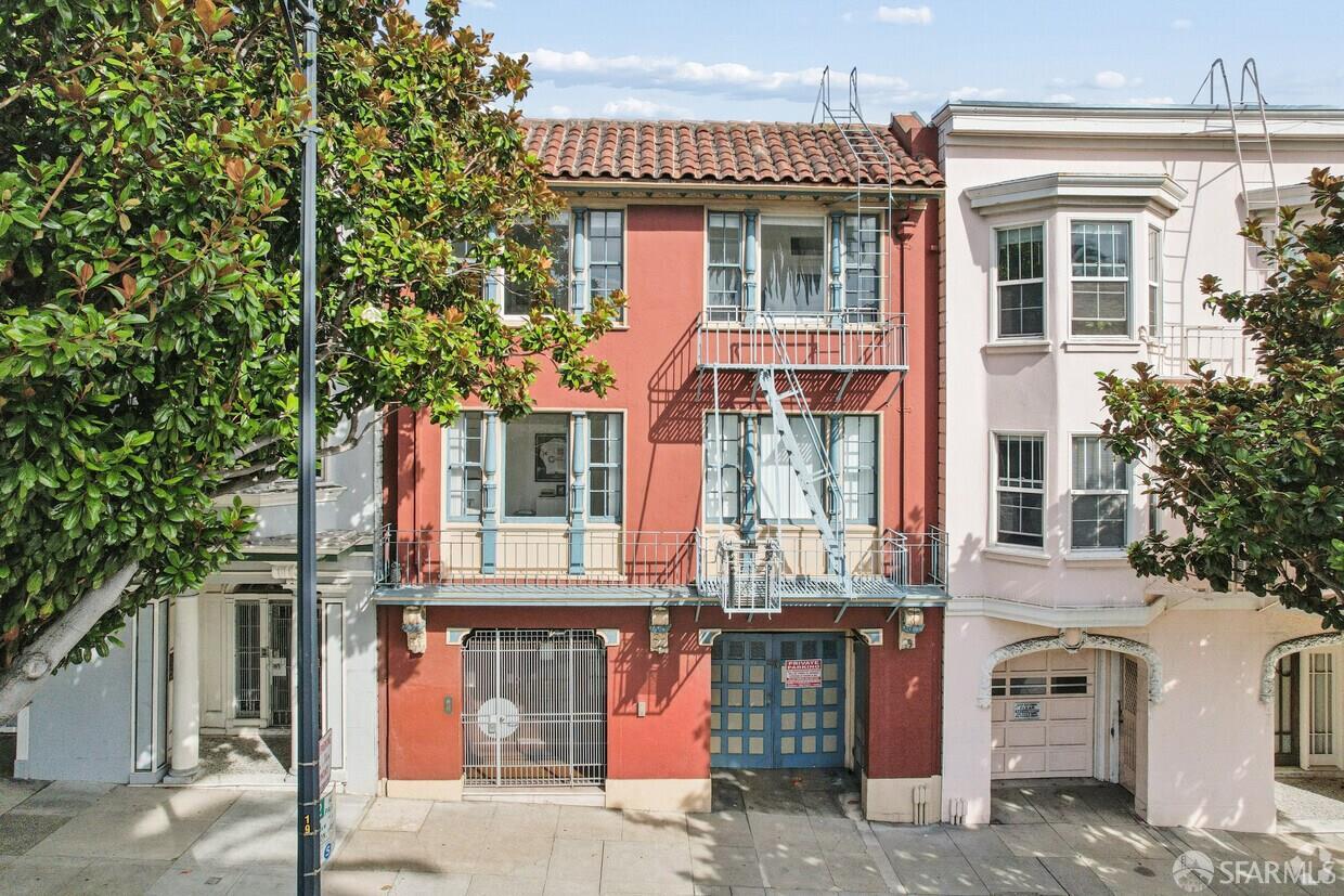 Red multi-story home with blue trim, balconies, and a metal fire escape, framed by leafy trees.