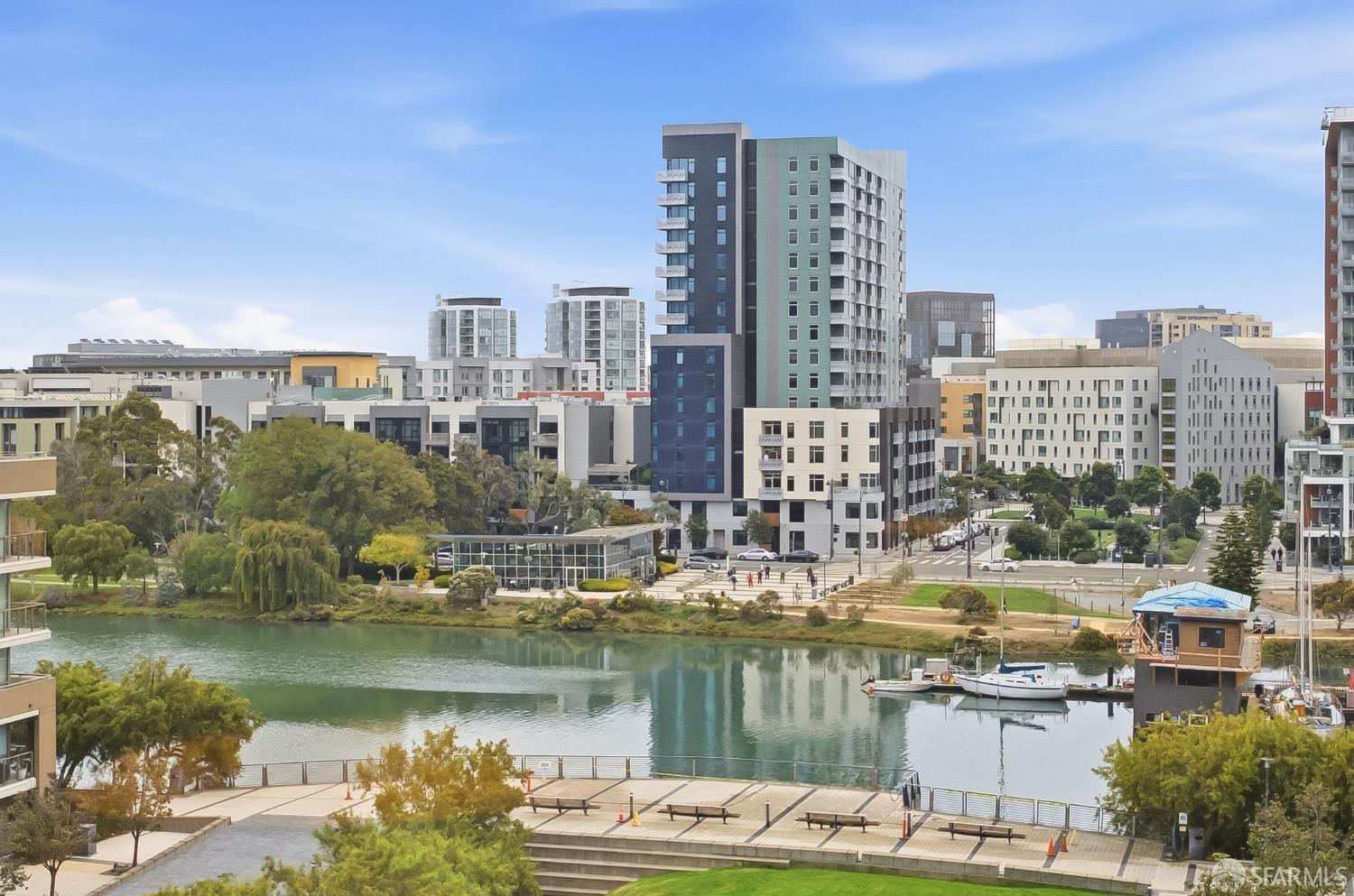 Urban waterfront with modern apartment buildings, boats, and a park promenade.