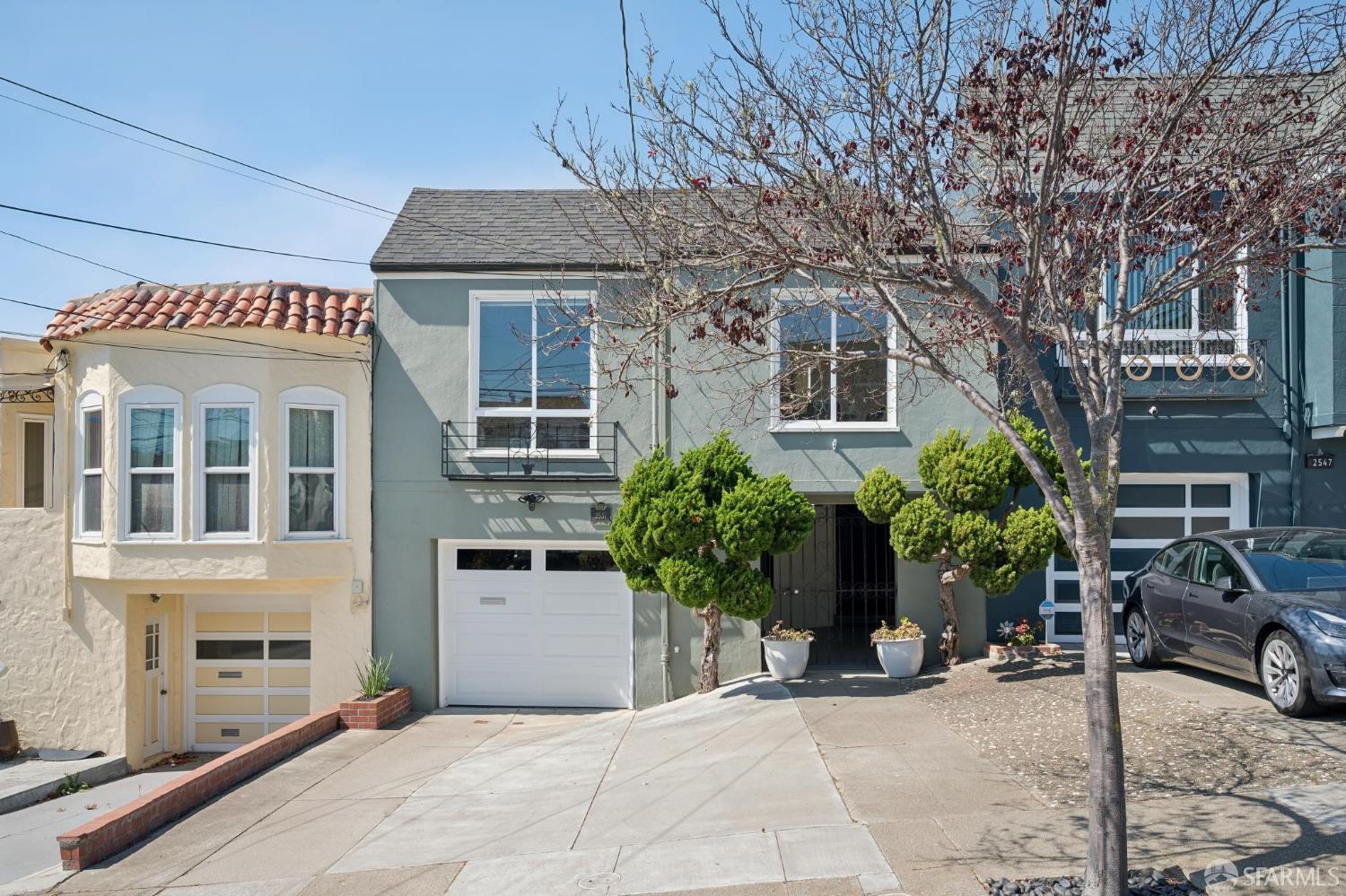 Sage-green two-story house with a small balcony, white garage door, and sculpted shrubs.