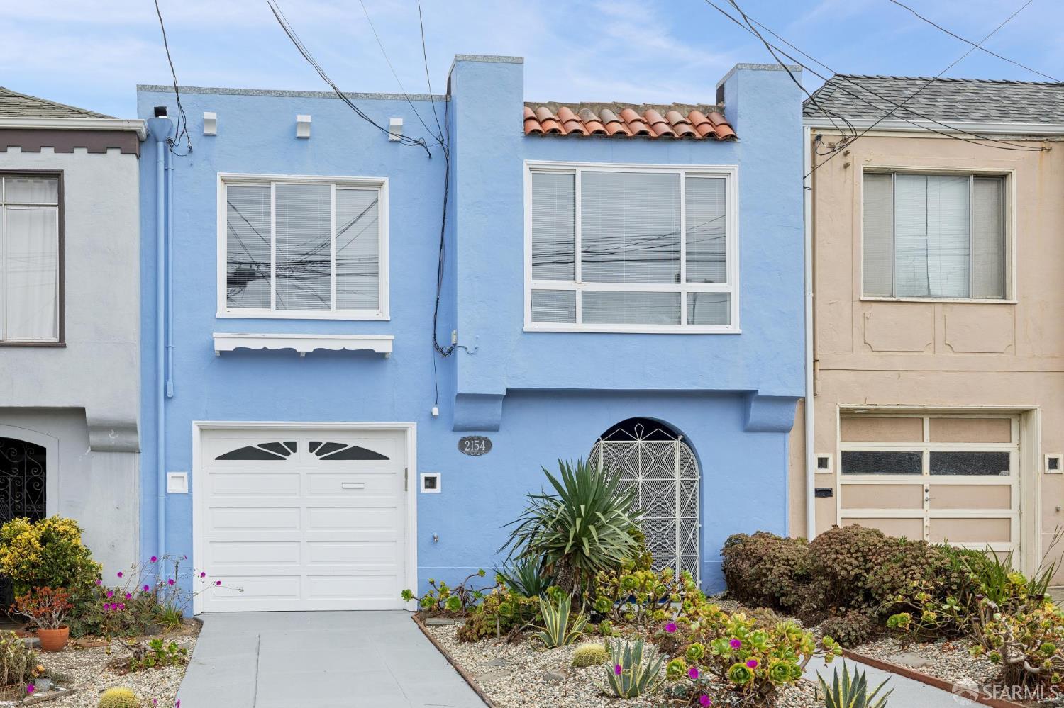 Blue two-story home with a white garage, arched security gate, and drought-tolerant front garden.