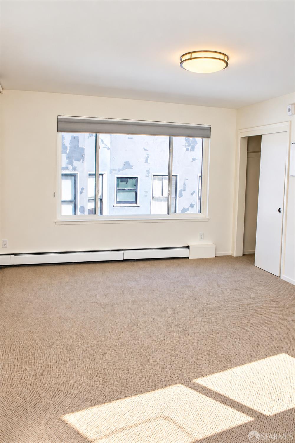 Empty carpeted bedroom with a wide window, sliding closet door, and sunlight across the floor.