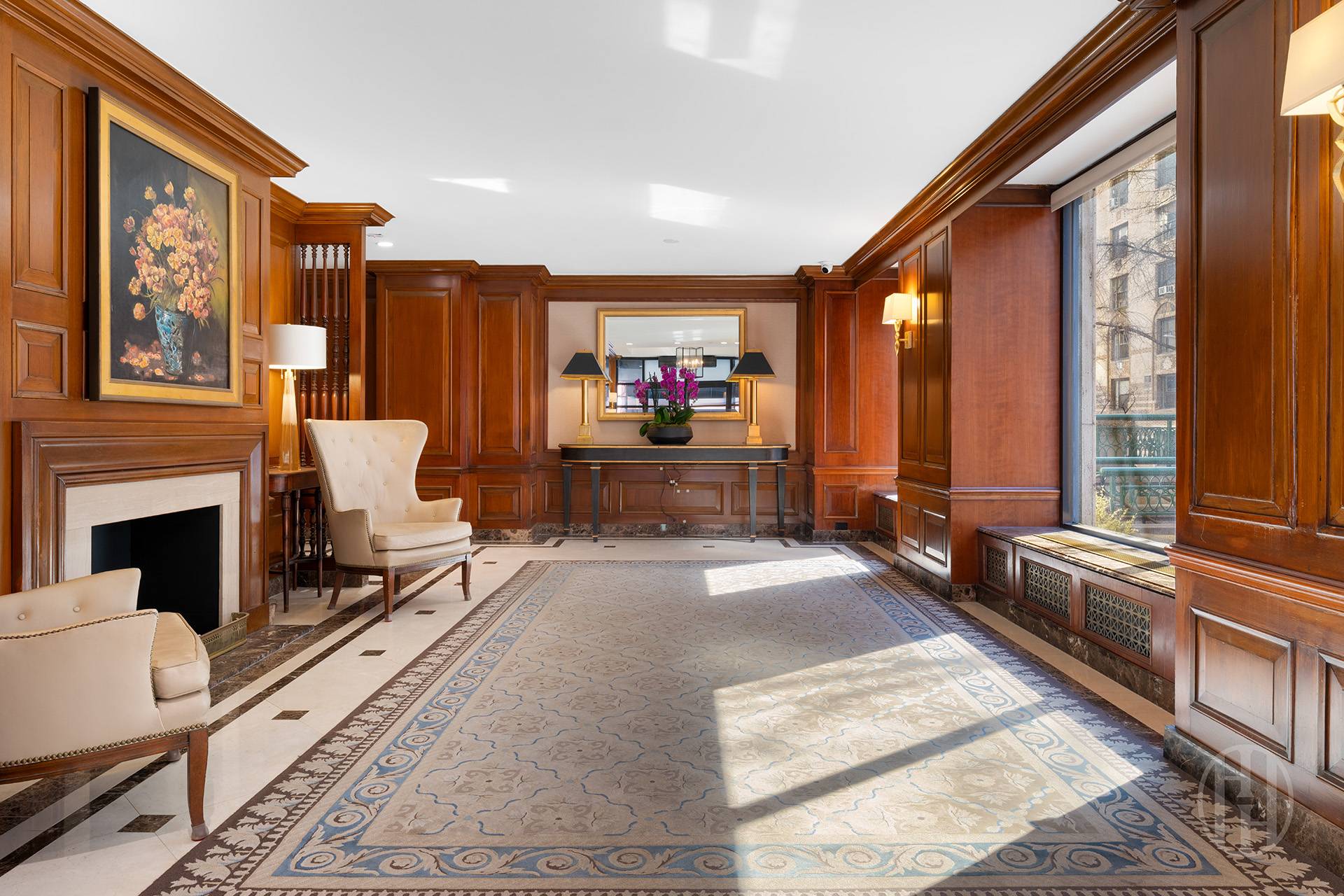 Wood-paneled sitting room with a fireplace, cream armchairs, patterned rug, and tall windows.