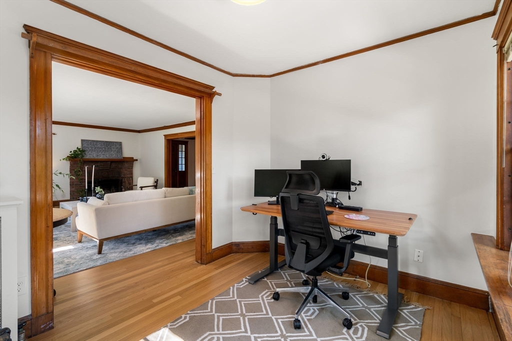 Home office with a standing desk and monitors, opening through a wood-trimmed doorway to a living room with a fireplace.