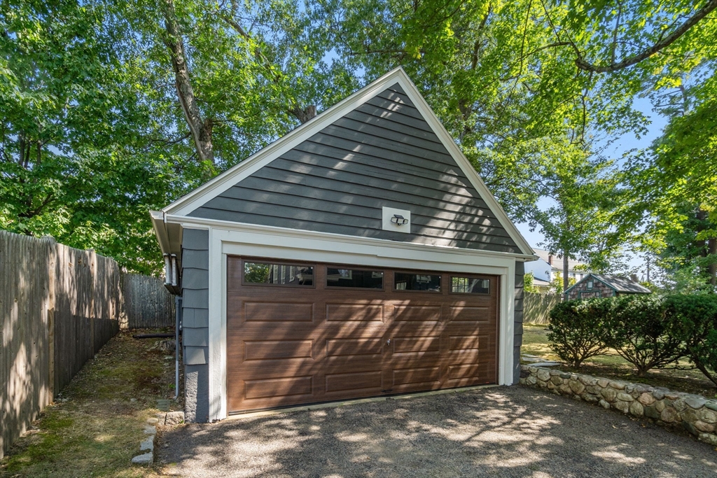 Detached gray garage with a brown double door, gravel driveway, and leafy trees.