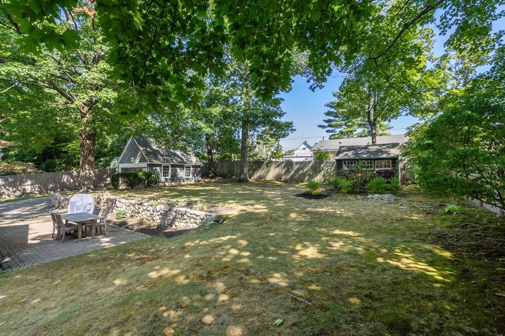 Shaded backyard with a paved patio, stone retaining wall, lawn, and two outbuildings.