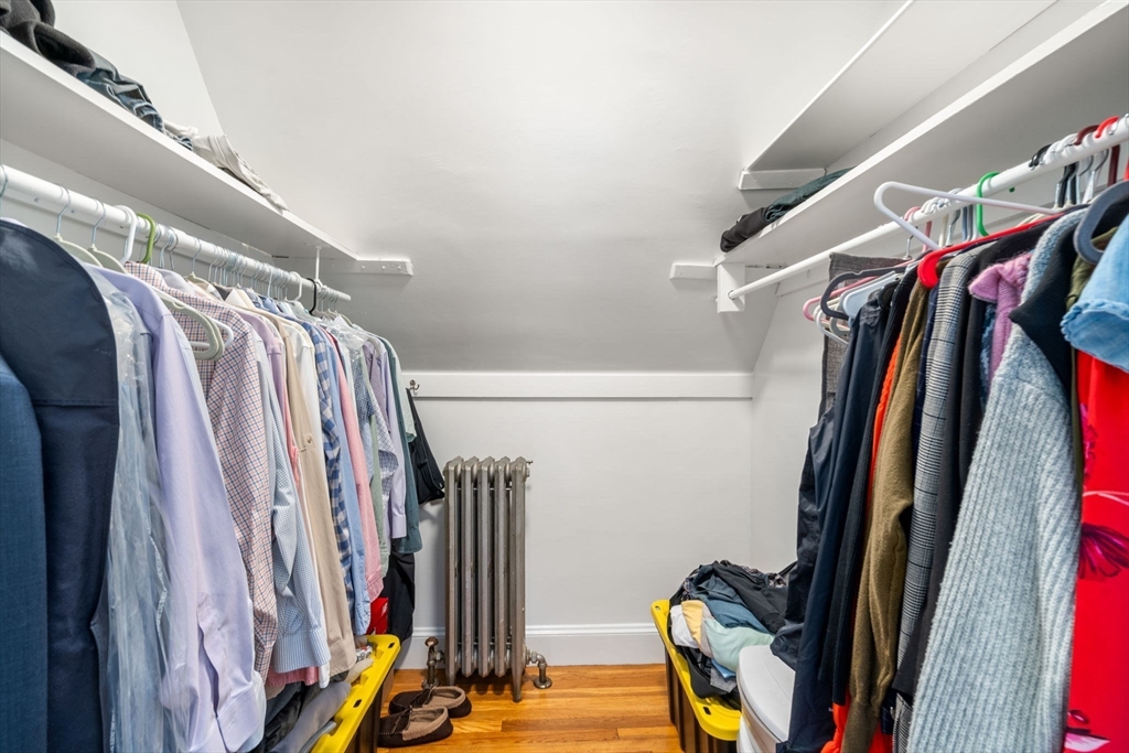 Walk-in closet with clothes on both sides, overhead shelves, a radiator, and hardwood flooring.