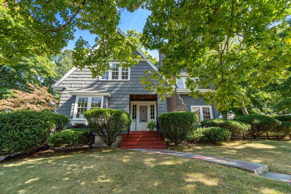 Front exterior of a gray shingle-sided home with red entry steps beneath leafy trees.