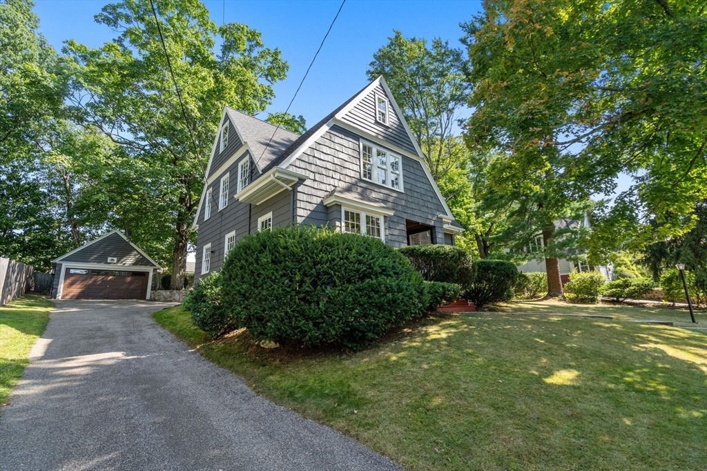 Gray shingle-sided house with steep gables, mature landscaping, and a detached garage beside the driveway.