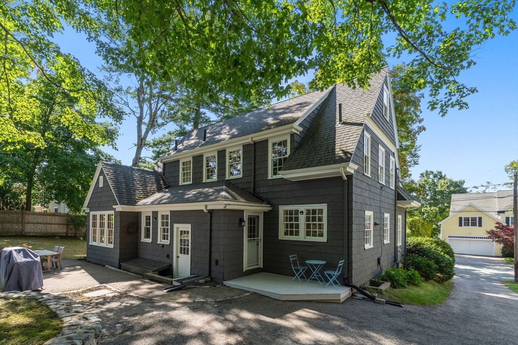Dark shingle house with white trim and a small patio with blue chairs beneath leafy trees.