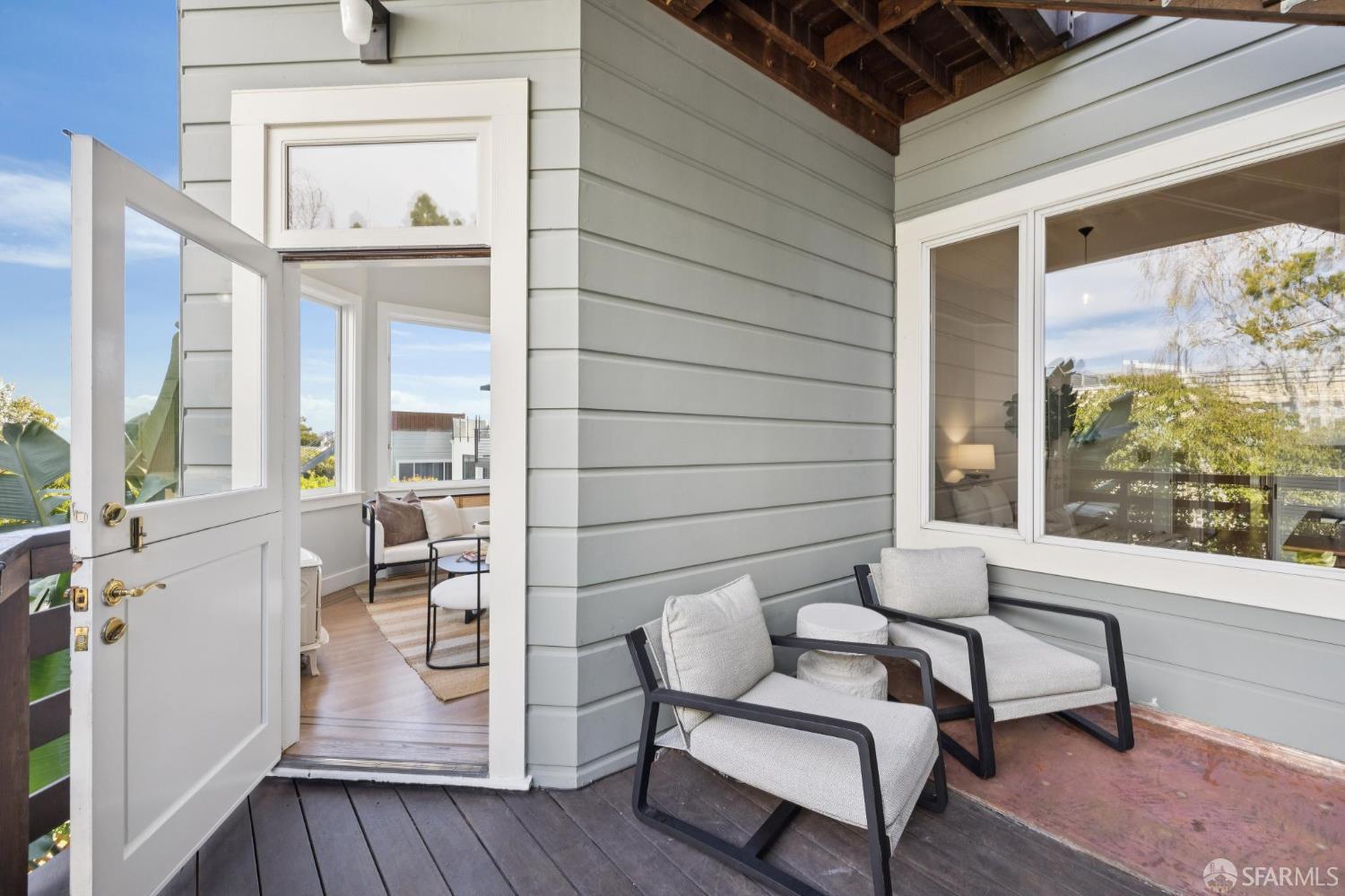 Covered deck with two cushioned chairs beside an open door to a bright room.