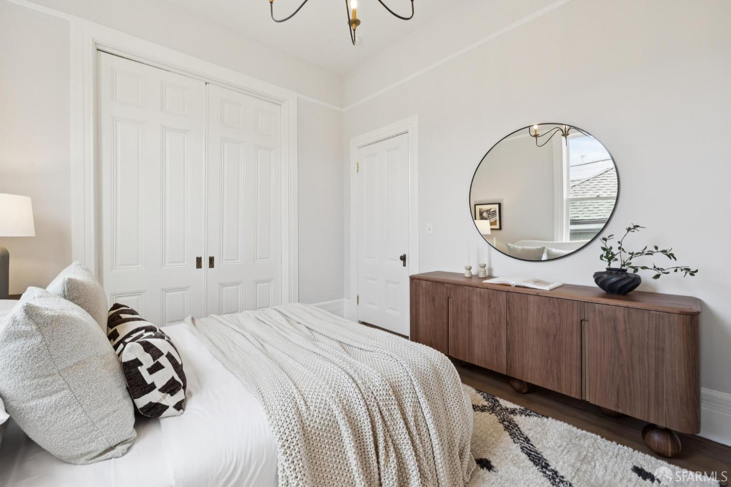 Staged bedroom with paneled closet doors, a round mirror, and a walnut sideboard.