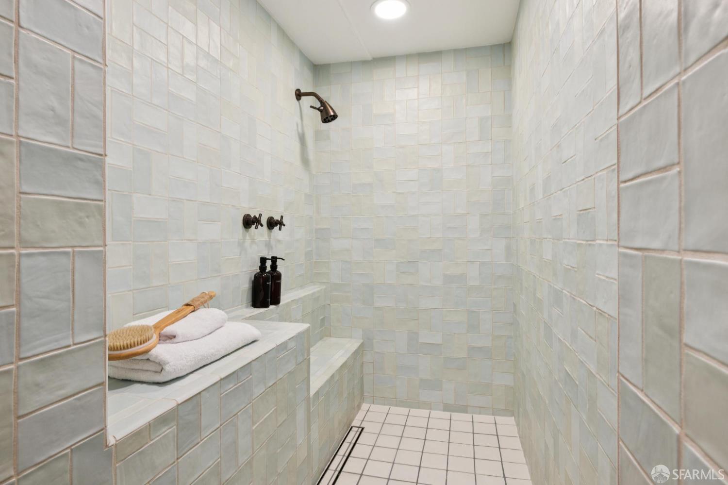 Walk-in shower with pale tiled walls, a built-in bench, folded towels, and bronze fixtures.