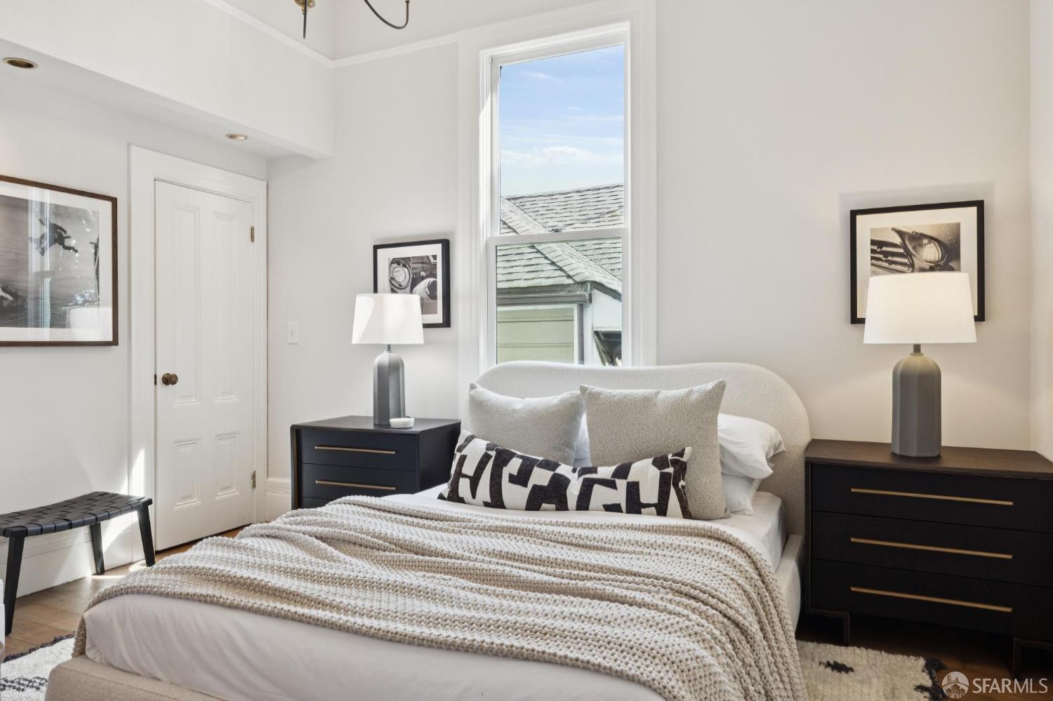 Staged bedroom with a cream upholstered bed, black bedside dressers, and a tall window.