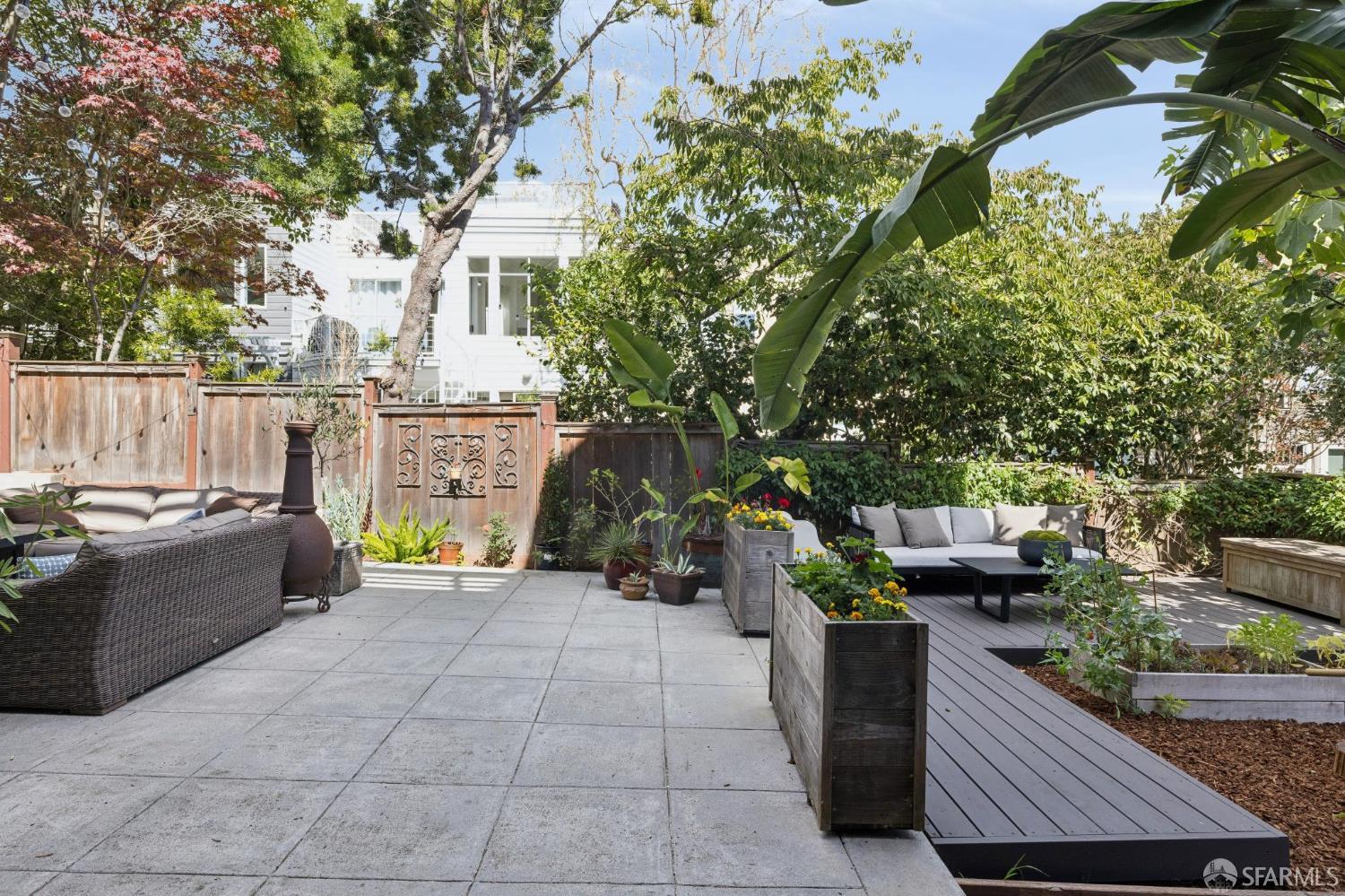 Landscaped backyard patio with paved terrace, raised deck, outdoor seating, and mature trees.
