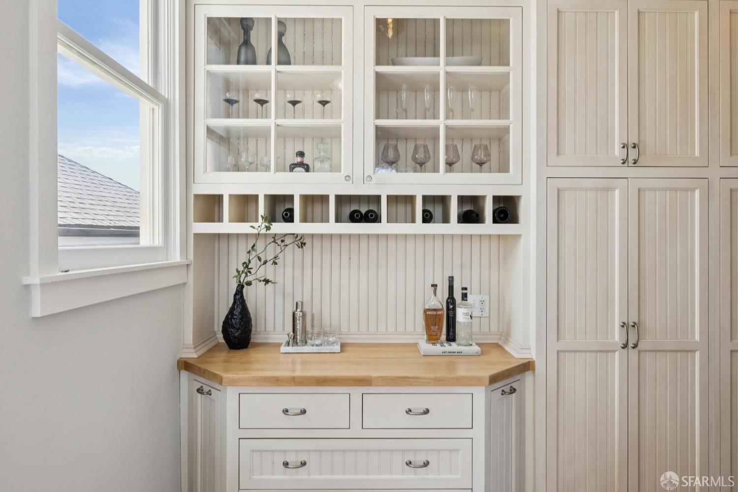 Built-in home bar with glass-front cabinets, displayed glassware, and a wood countertop beside a window.
