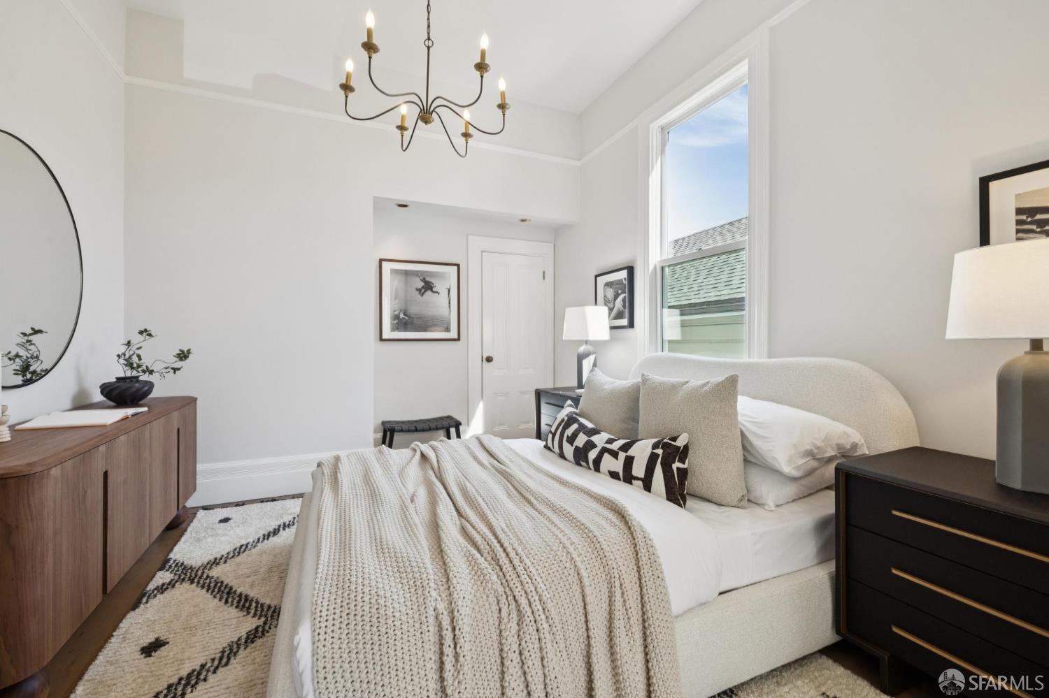 Staged bedroom with a white upholstered bed, cream bedding, tall window, and brass chandelier.