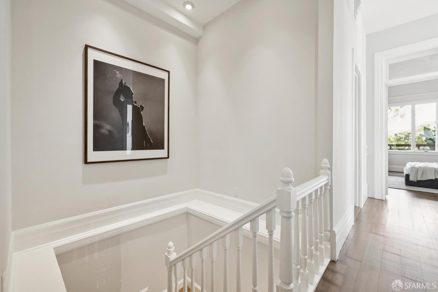 Upstairs hallway with a white stair railing, framed artwork, hardwood floors, and a sitting room beyond.