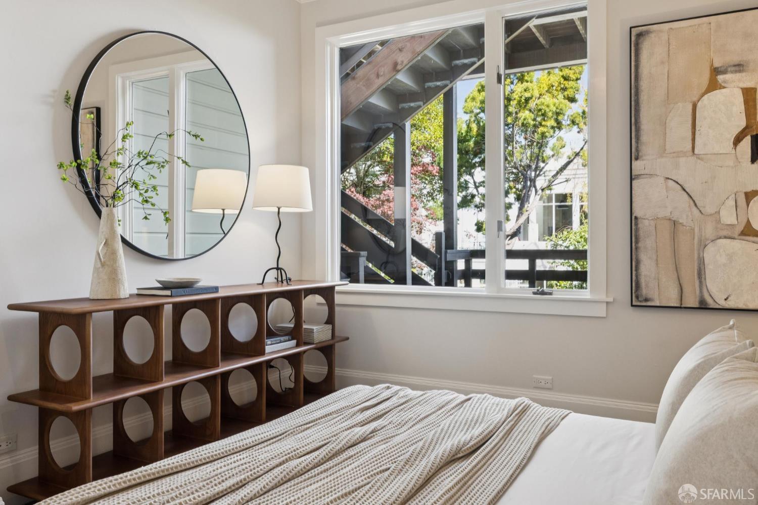 Bedroom with a dark wood console and round mirror beside large windows overlooking trees.