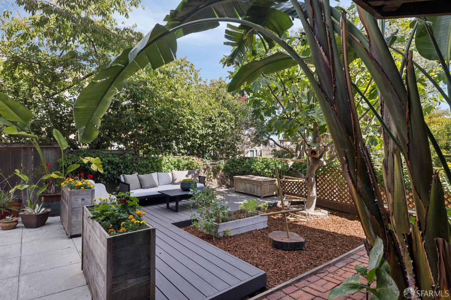 Backyard patio with a raised deck, outdoor seating, flower planters, and towering tropical foliage.