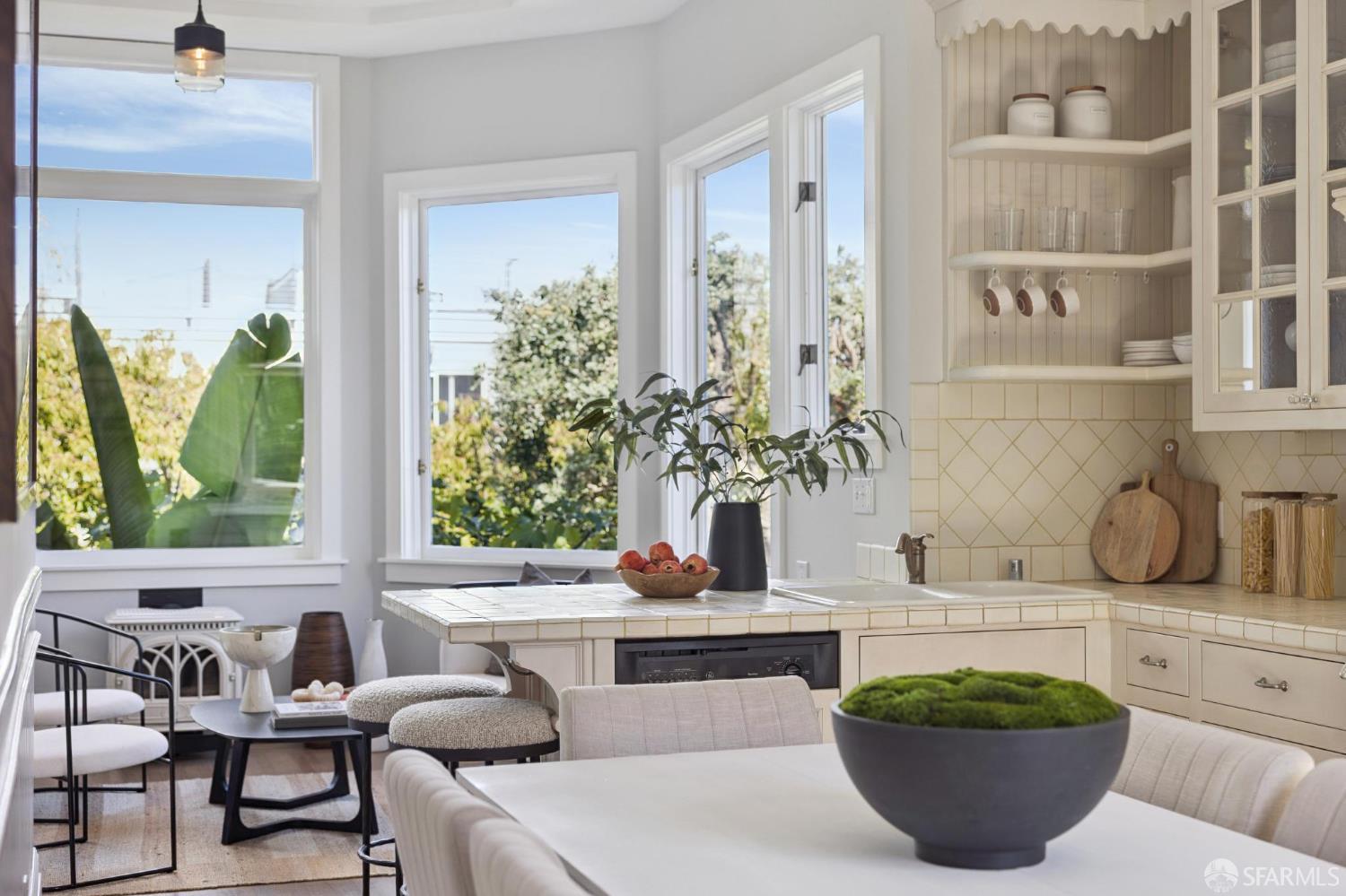Bright kitchen and dining area with cream tile counters, open shelves, and tall windows overlooking trees.