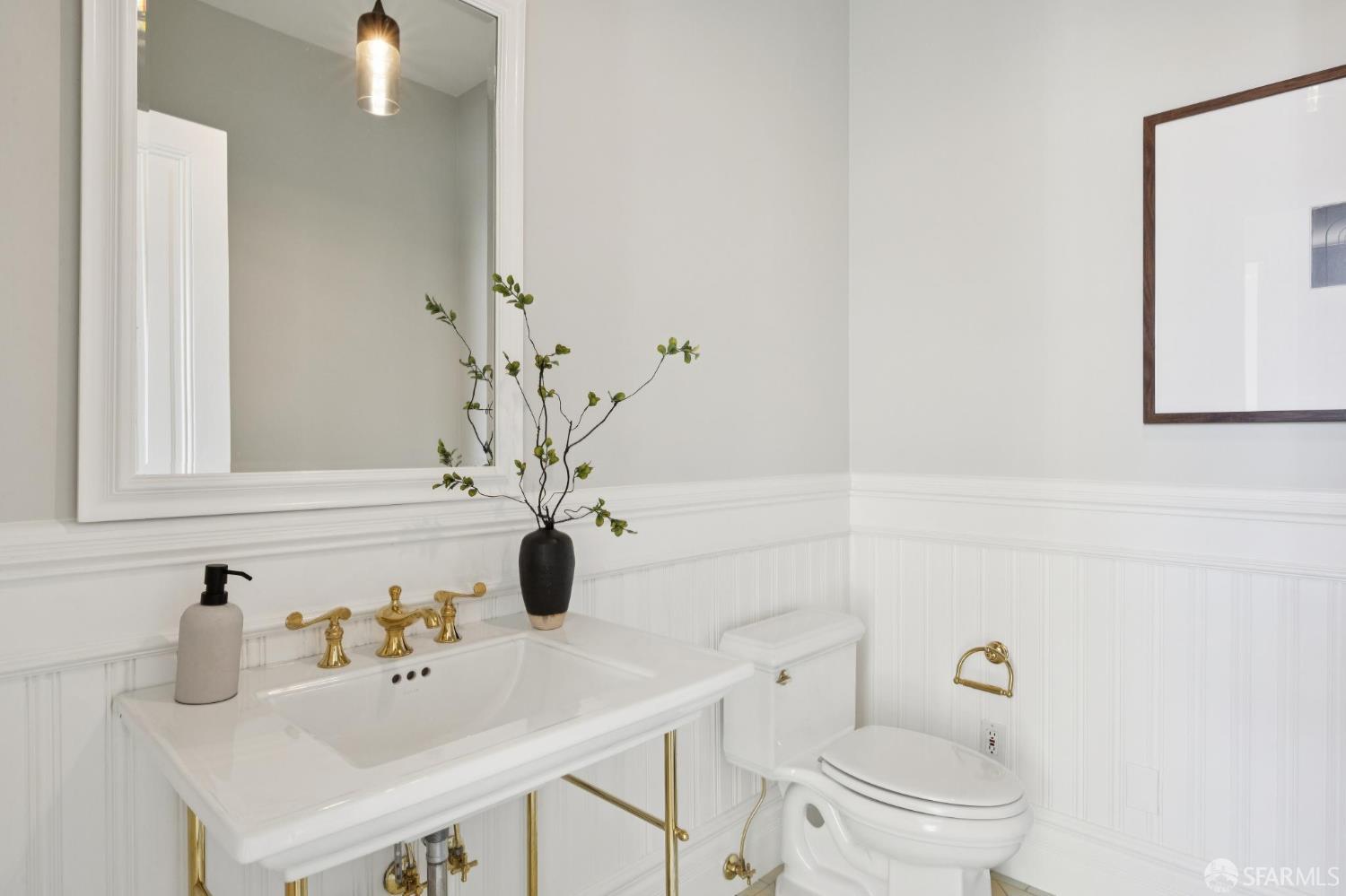Bright bathroom with a brass-framed sink, gold faucet, large mirror, and white beadboard wainscoting.