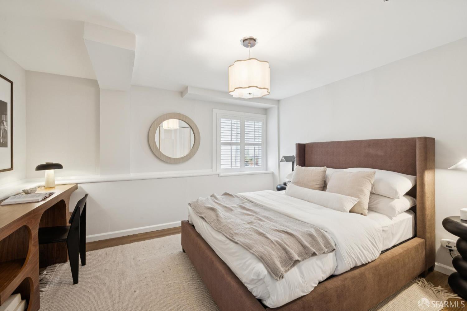 Furnished bedroom with a large bed, brown upholstered headboard, round mirror, and shuttered window.