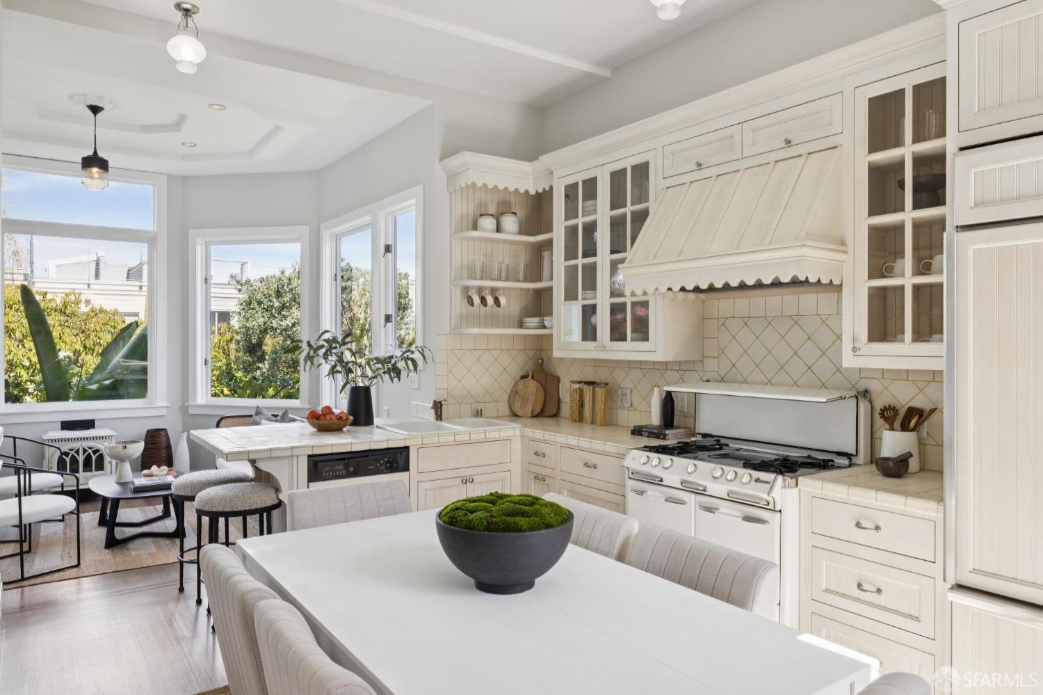 White kitchen with a large range, tiled counters, and a dining table beside bright windows.
