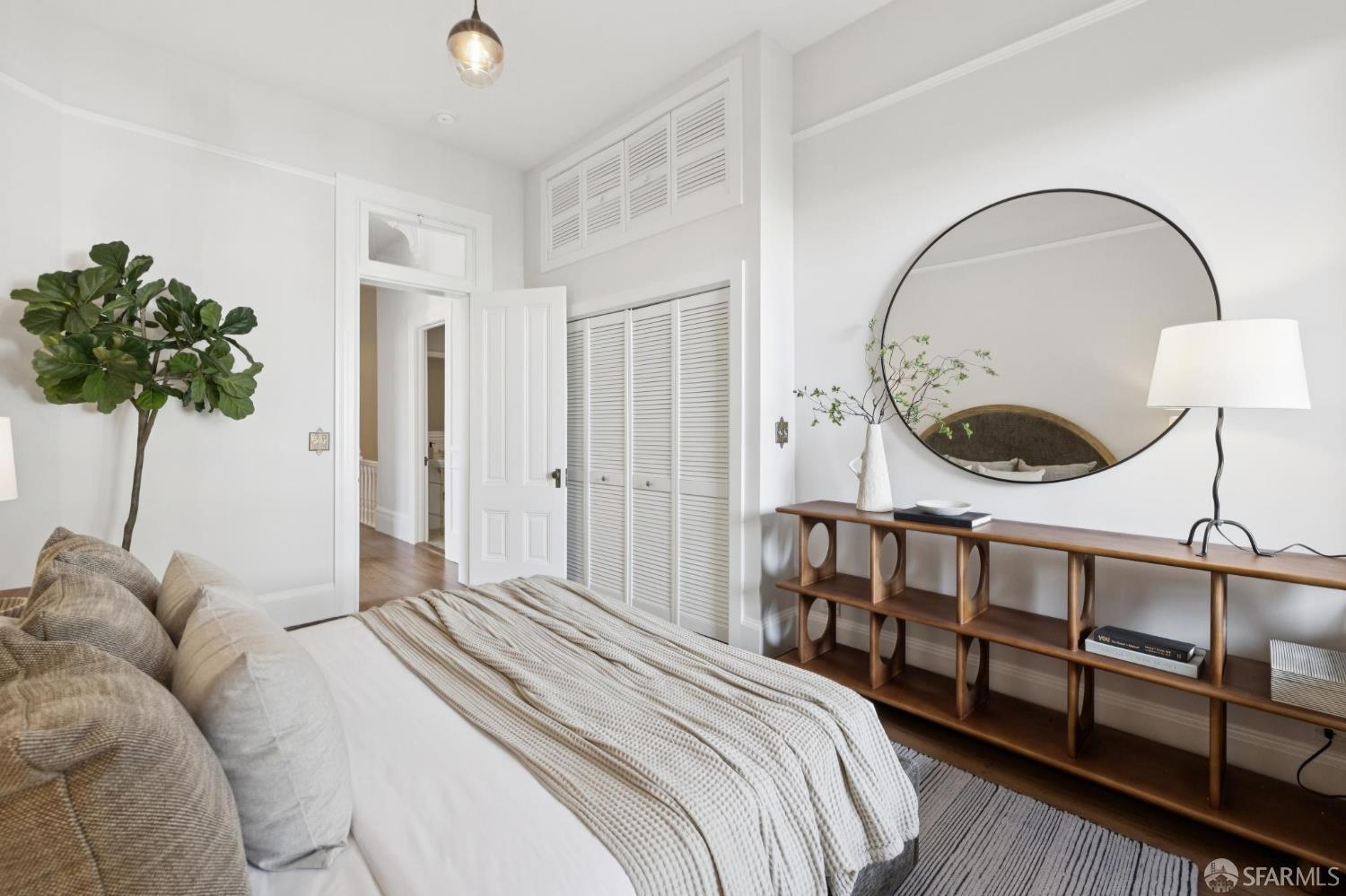Staged bedroom with white bedding, louvered closets, and a large round mirror.