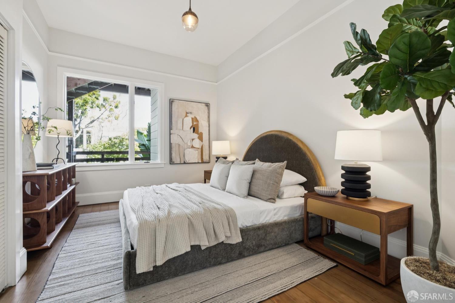 Staged bedroom with a curved wood-trimmed headboard, neutral bedding, and a large potted tree.