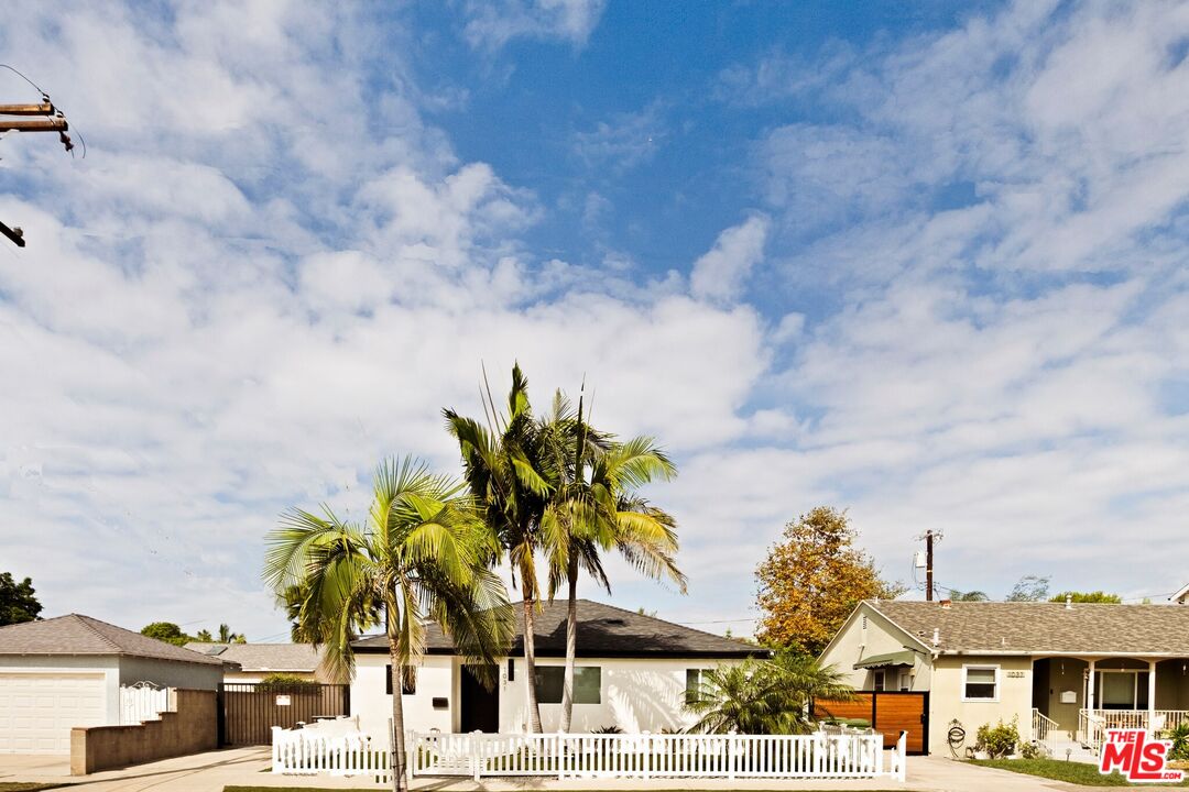 Single-story home behind a white picket fence, framed by palm trees and neighboring houses.