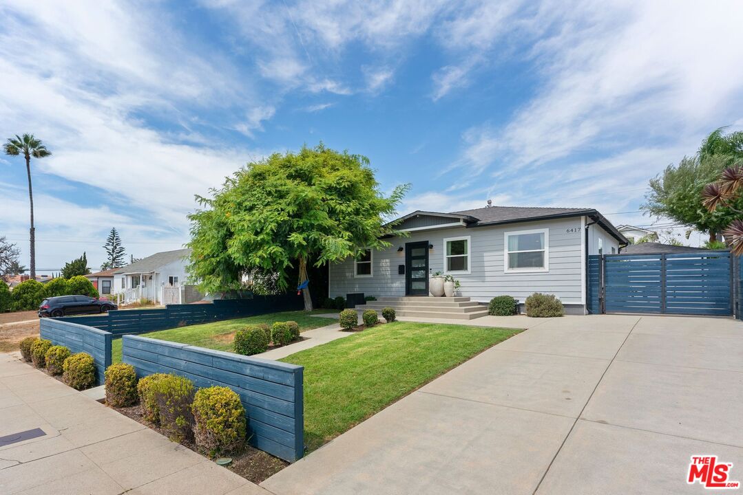 Single-story gray home with a broad driveway, blue fencing, and a landscaped front lawn.
